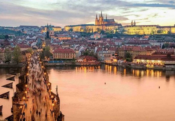 Widok na zachodzące słońce nad Pragą z widocznym Zamkiem Praskim i katedrą św. Wita w oddali, a na pierwszym planie tłum ludzi przechodzących przez kamienny Most Karola nad rzeką Wełtawą.