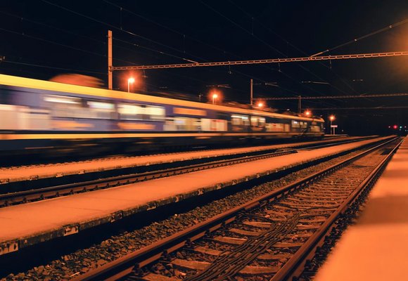 Pędzący pociąg na torach kolejowych nocą z rozmazanym efektem ruchu, oświetlony ulicznymi latarniami, z widoczną siecią trakcyjną i sygnalizacją świetlną w tle.