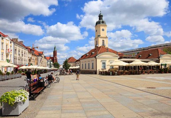 Widok na malowniczy, słoneczny rynek w Białymstoku z widocznym zegarem na wieży, idealne miejsce na spacer z kartą EKUZ w kieszeni. Na pierwszym planie ławki z siedzącymi ludźmi, rowery oraz kwiaty, a w tle restauracje z parasolami.