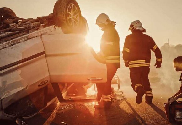 Wypadek na drodze, przewrócony biały samochód osobowy leży na boku na asfaltowej drodze podczas zachodu słońca. Strażacy w pełnym umundurowaniu podejmują działania ratunkowe