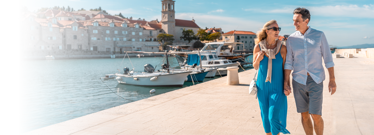 Dwie dorosłe osoby spacerują promenadą przy marinie z zacumowanymi łodziami na tle nadmorskiego miasteczka.