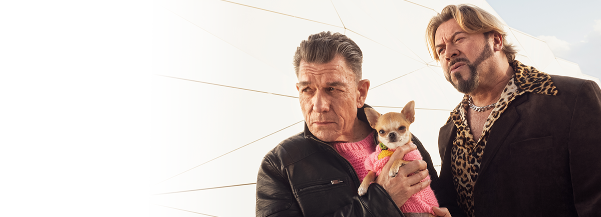 Dwie osoby stojące obok siebie, jedna trzymająca Chihuahua w różowym sweterku, obie w kurtkach skórzanych, na tle geometrycznych struktur na zewnątrz.