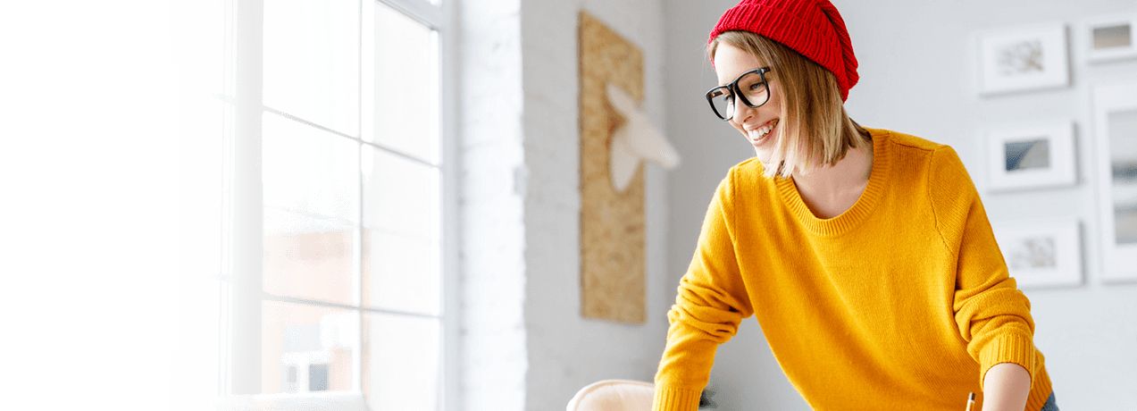 Uśmiechnięta kobieta ubrana w czerwoną czapkę, okulary i żółty sweter stoi w jasnym, nowoczesnym pokoju ze zdjęciami w ramkach na ścianie.