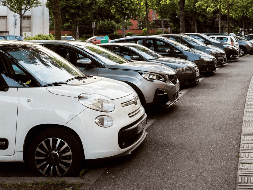 Zdjęcie przedstawia rząd zaparkowanych aut różnych marek wzdłuż ulicy, co może wiązać się z ryzykiem szkód parkingowych; pojazdy ustawione równolegle do chodnika, stwarzające typową scenę miejskiego parkowania.