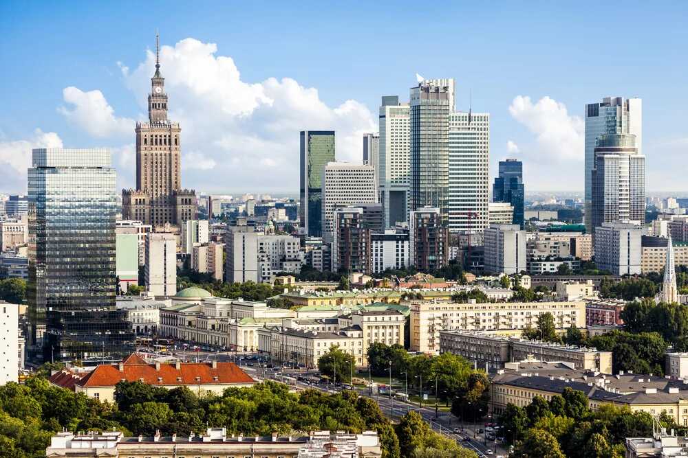 Widok na nowoczesne centrum Warszawy w słoneczny dzień, z charakterystycznym Pałacem Kultury i Nauki oraz otaczającymi go wysokimi budynkami biurowymi. Tło idealne do tematu podróży i korzystania z karty EKUZ podczas zagranicznych wizyt.