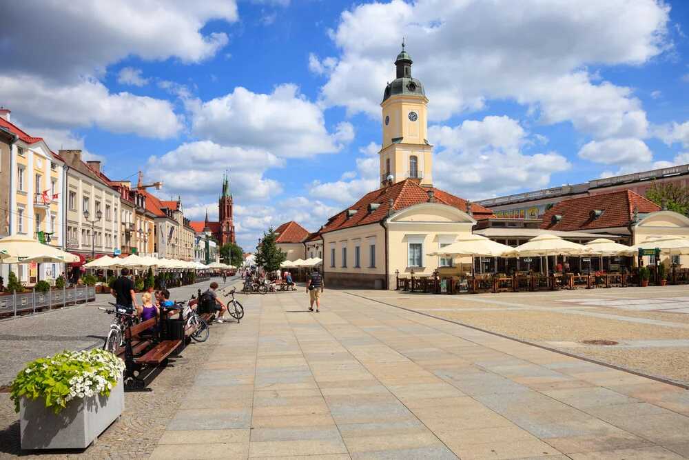 Widok na malowniczy, słoneczny rynek w Białymstoku z widocznym zegarem na wieży, idealne miejsce na spacer z kartą EKUZ w kieszeni. Na pierwszym planie ławki z siedzącymi ludźmi, rowery oraz kwiaty, a w tle restauracje z parasolami.