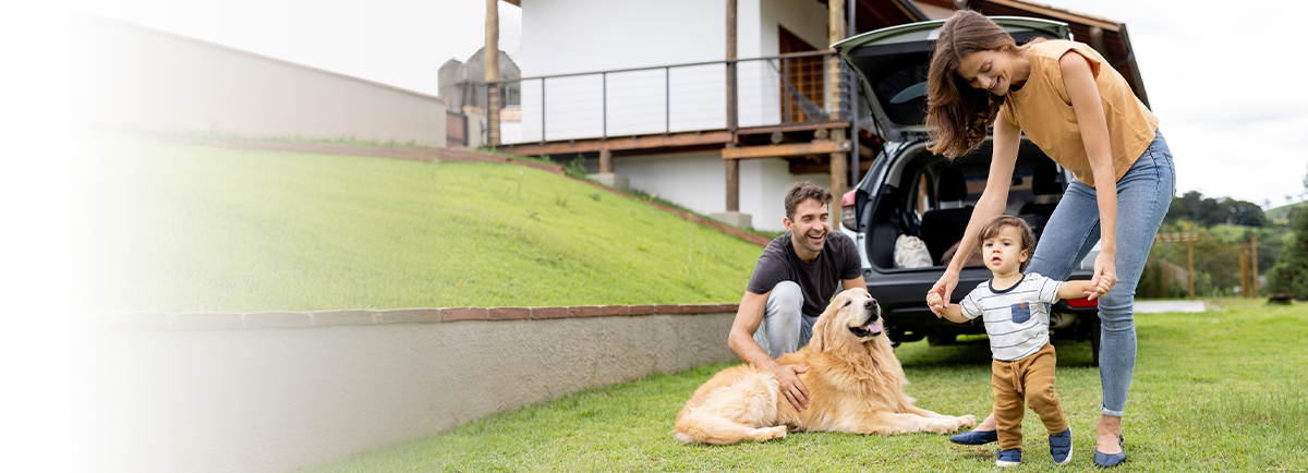 Trzy osoby i pies na trawie przed drewnianym domem, w tle otwarty bagażnik samochodu