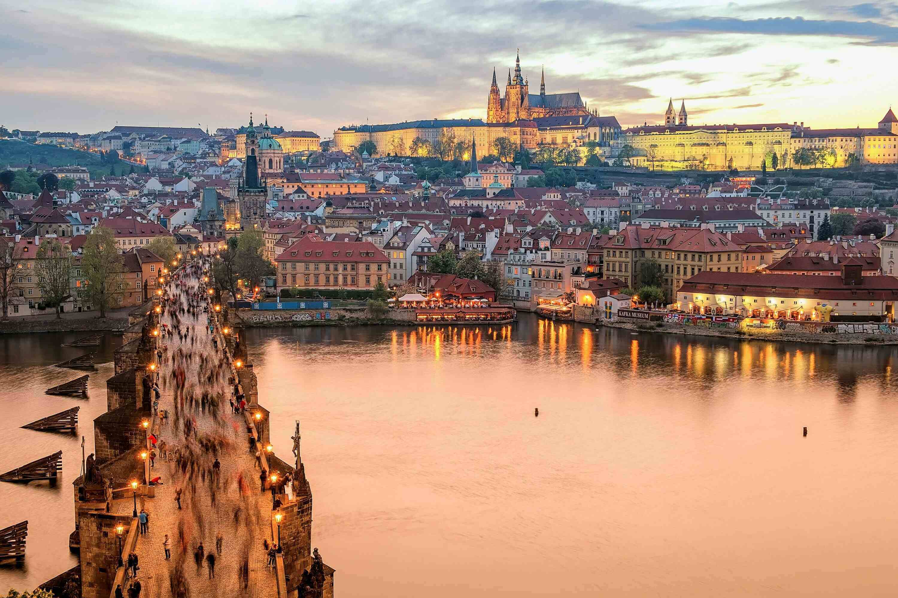 Widok na zachodzące słońce nad Pragą z widocznym Zamkiem Praskim i katedrą św. Wita w oddali, a na pierwszym planie tłum ludzi przechodzących przez kamienny Most Karola nad rzeką Wełtawą.